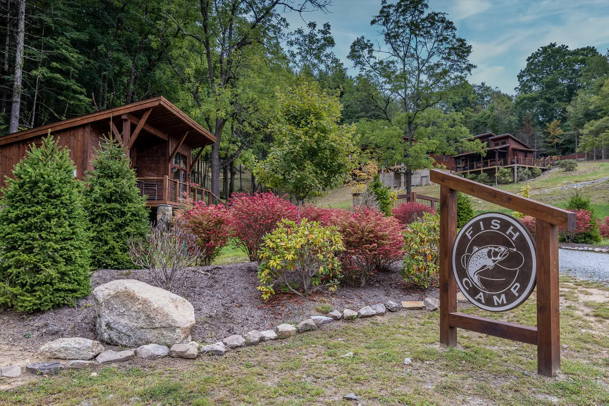 A collection of rustic wooden cabins set amidst landscaped gardens, with a 'Fish Camp' sign and plenty of outdoor space. The scene is lively yet relaxed, hinting at nearby activities like hiking and outdoor gatherings. The abundant greenery provides a sense of immersion in nature.