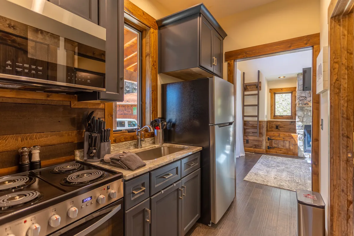 A sunlit kitchen with a prominent stove, sink, refrigerator, and windows flanked by wooden cabinetry. The space exudes warmth and functionality, with natural light enhancing the cozy, homey vibe. It feels ideal for both family gatherings and intimate meals, capturing the heart of culinary activity in the home.