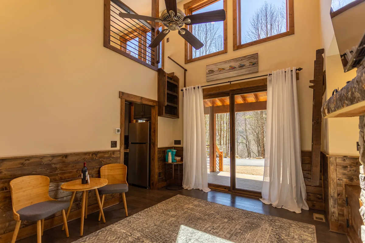 The living area features large windows that let in natural light, a stylish dining area, wooden furniture, and a stone fireplace. The atmosphere is both cozy and open, with space for both relaxation and entertaining guests. The fireplace, wooden accents, and view to the outdoors reinforce the space as warm and communal.