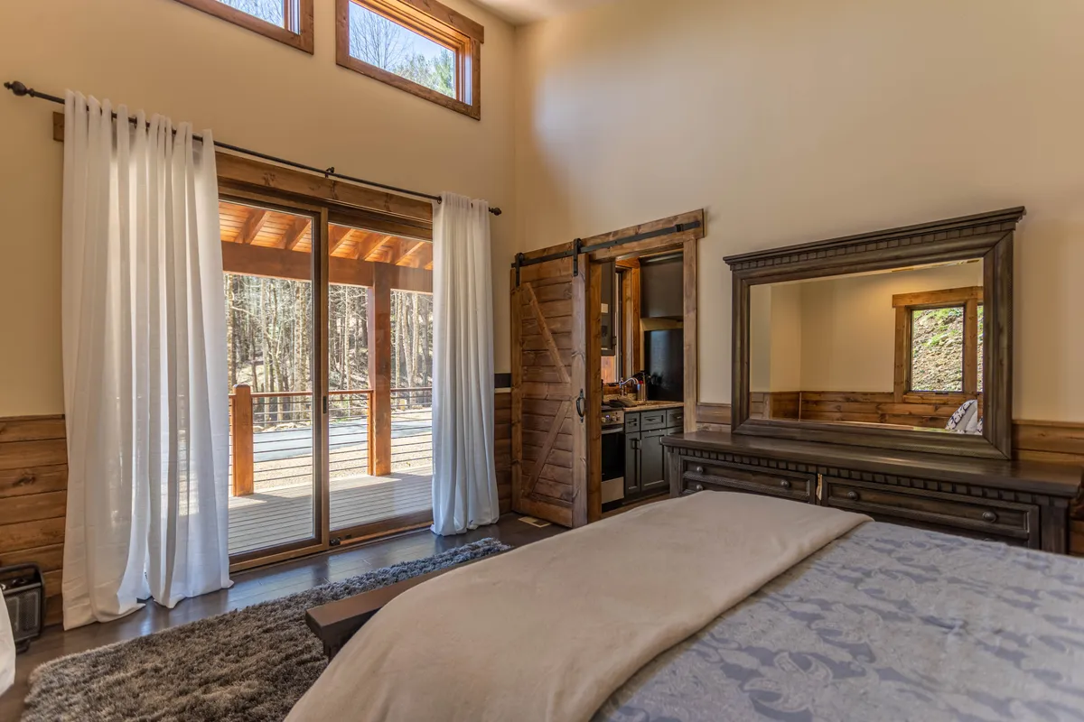 This image features a bed with decorative bedding, a large mirror reflecting natural light, sliding glass doors opening to the outdoors, and warm wooden accents throughout the room. The spacious layout and soft color palette create a calming, inviting bedroom atmosphere.