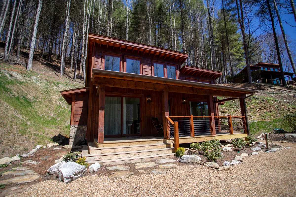 The hero image showcases a cozy wooden cabin exterior nestled among lush greenery with large windows reflecting the natural surroundings. A welcoming front porch, complete with comfortable seating, invites guests to relax and savor the tranquil ambiance. A landscaped pathway winds toward the entrance, hinting at serene nature walks right outside the door.