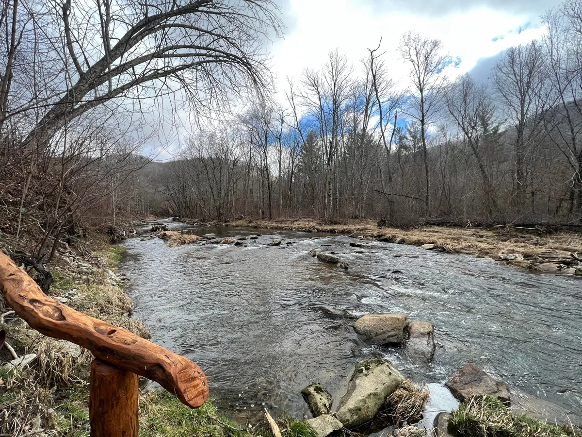 A tranquil scene featuring a flowing river bordered by weathered rocks and a wooden railing, with wildflower blooms in spring and rich sunset reflections on the water. The surrounding area is perfect for activities such as fishing, picnicking, hiking, and wildlife observation, offering a vibrant yet peaceful natural setting.