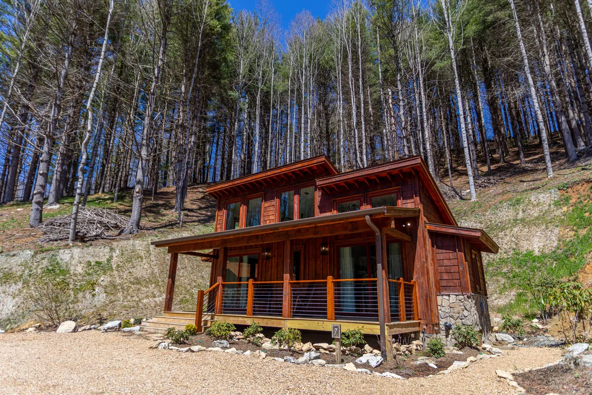 A charming wooden cabin nestled amidst tall trees, featuring a welcoming porch perfect for relaxing. The setting exudes tranquility and is surrounded by nature, suggesting privacy and a cozy retreat. The porch area hints at possibilities for enjoying morning coffee or romantic evenings outdoors.
