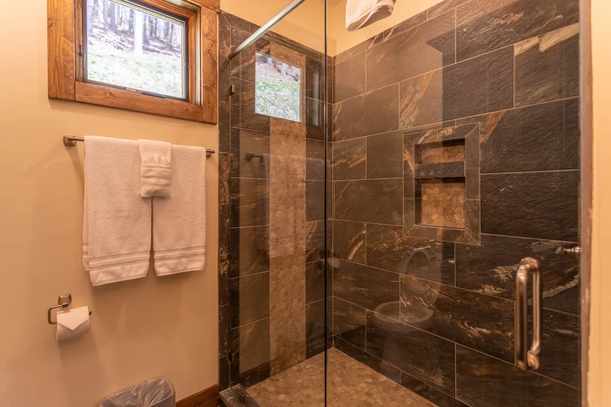 The bathroom showcases a glass shower enclosure with beautifully textured stone tiles and wooden window frames, creating a spa-like feel. The space is designed for relaxation, featuring a refreshing walk-in shower and a granite vanity that adds an element of luxury.